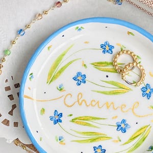 May include: A white ceramic trinket dish with a blue rim. The dish is decorated with a floral pattern of blue forget-me-not flowers and green leaves. The word "Chanel" is written in gold script on the dish. Two gold rings are sitting on the dish.