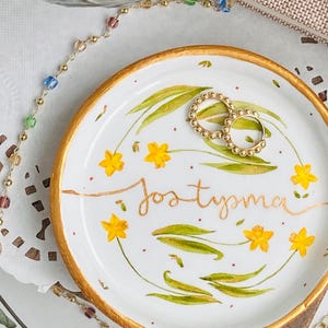 May include: A white ceramic ring dish with a gold rim and a floral design featuring yellow daffodils and green leaves. The dish has the word "Joslynma" written in gold script. Two gold rings are resting on the dish.