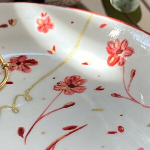 May include: A white ceramic trinket dish with a red floral pattern and gold accents. The dish is perfect for holding jewelry, keys, or other small items.
