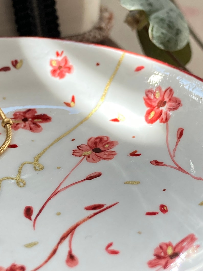 May include: A white ceramic trinket dish with a red floral pattern and gold accents. The dish is perfect for holding jewelry, keys, or other small items.
