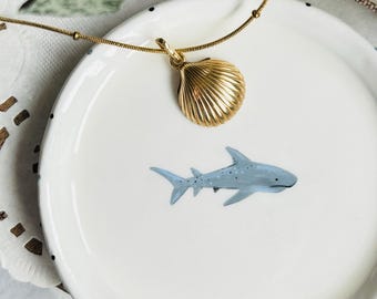 May include: A gold chain necklace with a seashell pendant. The necklace is laying on a white ceramic dish with a blue shark illustration.