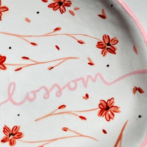 May include: White ceramic dish with a pink rim and a floral design. The dish has a pink inscription that reads "blossom".