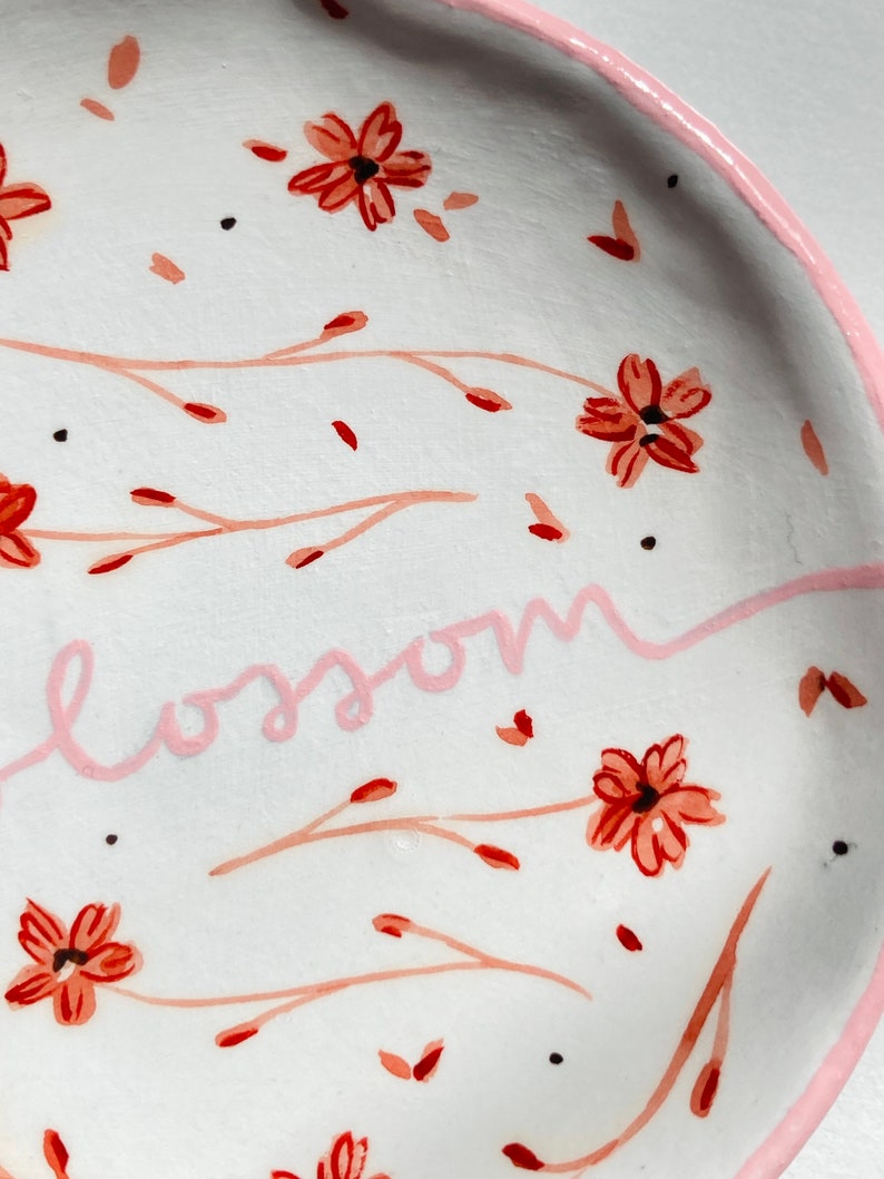 May include: White ceramic dish with a pink rim and a floral design. The dish has a pink inscription that reads "blossom".