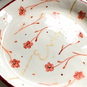 May include: A white ceramic dish with a red rim and a floral design. The dish is decorated with small red flowers and gold lines.