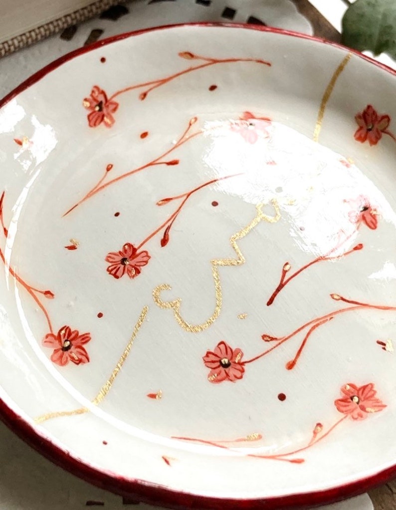May include: A white ceramic dish with a red rim and a floral design. The dish is decorated with small red flowers and gold lines.