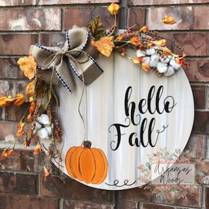 May include: A round wooden door hanger with the words "hello fall" in black script. The hanger is decorated with a burlap bow, fall leaves, cotton bolls, and a painted orange pumpkin.
