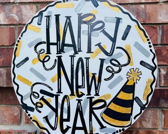 May include: Round wooden sign with the words "HAPPY NEW YEAR" in black, with a party hat and confetti-like shapes in gold, silver, and black. The sign is white with a black border and is hanging on a brick wall.