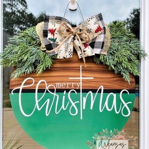 May include: A round wooden door hanger with a green and brown woodgrain design. The top half of the door hanger is brown woodgrain and the bottom half is green. The words "merry christmas" are written in white cursive lettering on the green portion of the door hanger. A white cross is centered above the word "merry". The door hanger is decorated with a large bow made of black and white buffalo plaid fabric and a cream fabric with red and green Christmas trees. The bow is tied with twine. The door hanger is hanging on a white hook. The words "Arkansas Woods Craft Co." are written in white lettering on a floral design in the bottom right corner of the image.