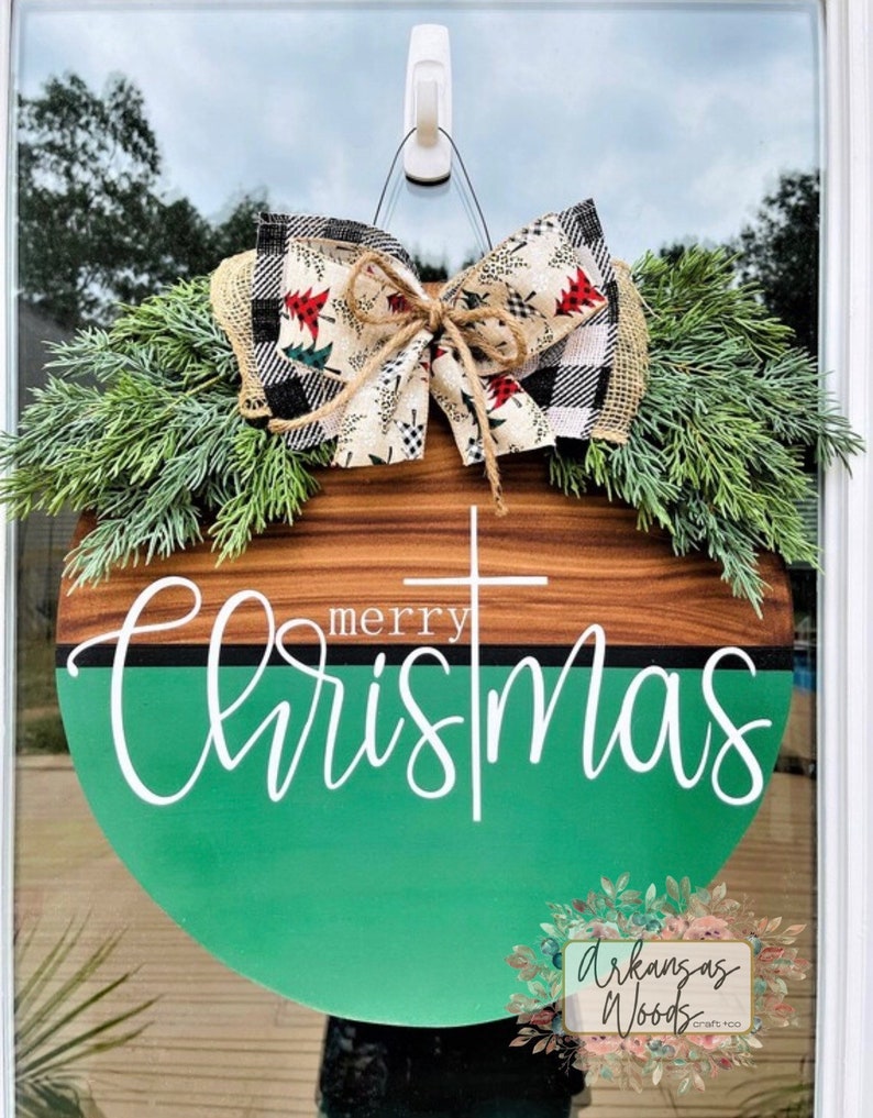 May include: A round wooden door hanger with a green and brown woodgrain design. The top half of the door hanger is brown woodgrain and the bottom half is green. The words "merry christmas" are written in white cursive lettering on the green portion of the door hanger. A white cross is centered above the word "merry". The door hanger is decorated with a large bow made of black and white buffalo plaid fabric and a cream fabric with red and green Christmas trees. The bow is tied with twine. The door hanger is hanging on a white hook. The words "Arkansas Woods Craft Co." are written in white lettering on a floral design in the bottom right corner of the image.