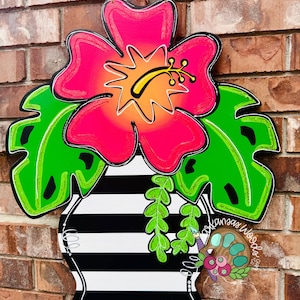 May include: A pink hibiscus flower with a yellow center in a black and white striped vase with green leaves. The vase is hanging on a brick wall.