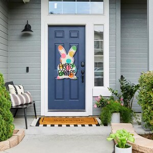 May include: A navy blue front door with a colorful Easter bunny decoration that reads "Happy Easter." The door is framed by a white trim and a window above. A welcome mat and potted plants are in front of the door.