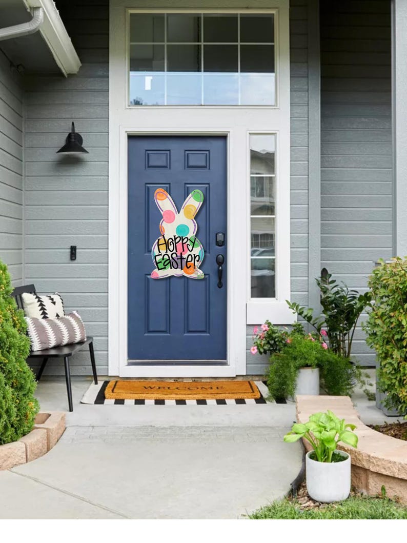 May include: A navy blue front door with a colorful Easter bunny decoration that reads "Happy Easter." The door is framed by a white trim and a window above. A welcome mat and potted plants are in front of the door.
