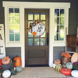 May include: A front porch decorated for fall with a wooden sign that says "FALL", a wreath with the text "hello fall", and pumpkins in various sizes and colors.