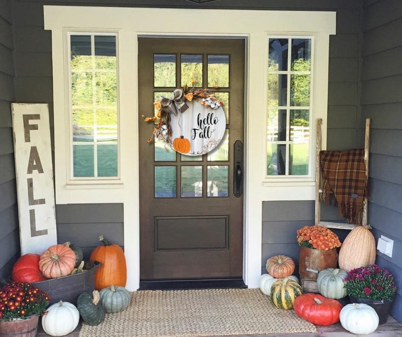 May include: A front porch decorated for fall with a wooden sign that says "FALL", a wreath with the text "hello fall", and pumpkins in various sizes and colors.