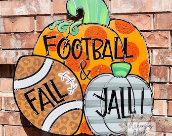 May include: A wooden door hanger shaped like a pumpkin with the words "Football & Fall Y'all" in black lettering. The pumpkin is orange with white polka dots and a green stem. A football with a brown and black leopard print is on the left side of the pumpkin. A smaller white pumpkin with gray stripes is on the right side of the pumpkin.