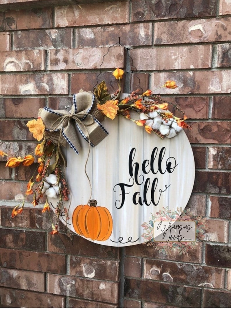 May include: A round wooden door hanger with the words "hello fall" in black script. The hanger is decorated with a burlap bow, fall leaves, cotton bolls, and a painted orange pumpkin.
