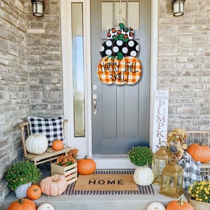 Peut inclure: Une couronne de porte sur le th&egrave;me de l'automne avec trois citrouilles empil&eacute;es. Les citrouilles sont peintes en orange, noir et blanc avec un motif &agrave; carreaux. La citrouille du haut a une tige verte et la citrouille du milieu porte l'inscription "Happy Fall Y'all". La couronne est accroch&eacute;e &agrave; une porte grise avec un paillasson noir qui dit "Feels Good To Be Home".