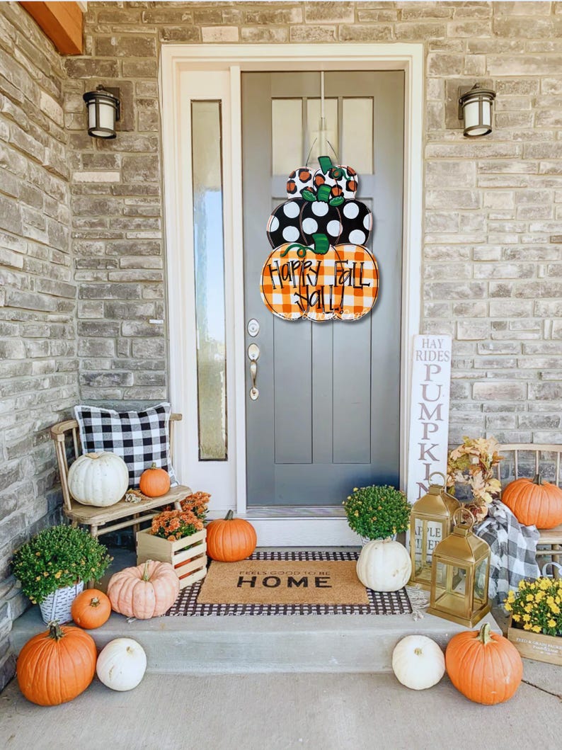 Peut inclure: Une couronne de porte sur le th&egrave;me de l'automne avec trois citrouilles empil&eacute;es. Les citrouilles sont peintes en orange, noir et blanc avec un motif &agrave; carreaux. La citrouille du haut a une tige verte et la citrouille du milieu porte l'inscription "Happy Fall Y'all". La couronne est accroch&eacute;e &agrave; une porte grise avec un paillasson noir qui dit "Feels Good To Be Home".