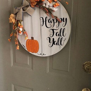 May include: A round wooden door hanger with a whitewashed background and black lettering that reads "Happy Fall Y'all". The hanger is decorated with a painted orange pumpkin, a brown burlap bow, and a floral arrangement of white cotton, orange flowers, and green leaves.
