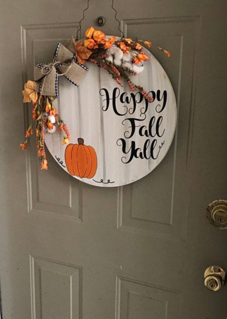 May include: A round wooden door hanger with a whitewashed background and black lettering that reads "Happy Fall Y'all". The hanger is decorated with a painted orange pumpkin, a brown burlap bow, and a floral arrangement of white cotton, orange flowers, and green leaves.