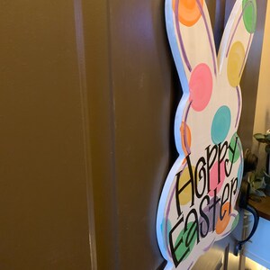 May include: A white wooden Easter bunny-shaped door hanger with colorful polka dots and the words "Hoppy Easter" in black script. The bunny has a painted outline and is hanging on a brown door.