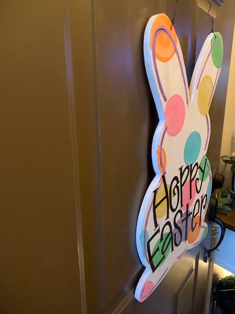 May include: A white wooden Easter bunny-shaped door hanger with colorful polka dots and the words "Hoppy Easter" in black script. The bunny has a painted outline and is hanging on a brown door.