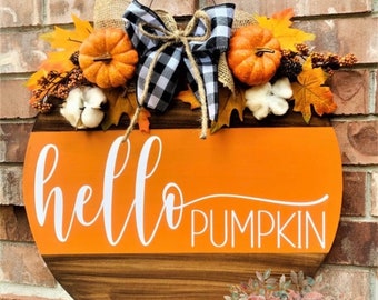 May include: A round wooden door hanger with a painted orange pumpkin design. The words "hello pumpkin" are written in white script on the orange pumpkin. The hanger is decorated with a burlap bow, a black and white checkered bow, and fall foliage.