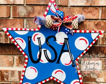 May include: A blue star-shaped wooden door hanger with a red, white, and blue striped border. The star has white polka dots and the word "USA" written in black. A red, white, and blue bow is attached to the top of the star.