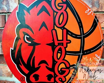 May include: A round wooden sign with a red and orange background. The left side features a black and red cartoon hog with the text "GO HOGS" in black and red on the right side. The sign has a basketball design on the right side.