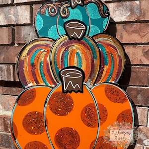 May include: A wooden pumpkin decoration with three stacked sections. The top section is teal with white polka dots, the middle section has orange, gold, and teal brushstrokes, and the bottom section is orange with large brown glitter dots. A metal wire is attached for hanging.