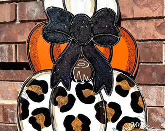 May include: A stacked pumpkin-shaped wooden door hanger with a black glitter bow. The top pumpkin is white, the middle is orange, and the bottom has a leopard print design. The hanger is against a brick wall.