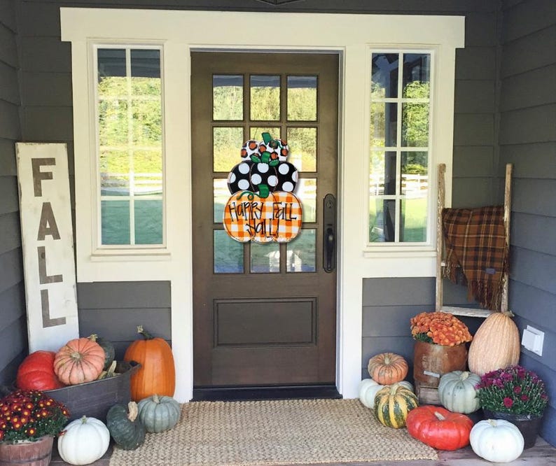 Peut inclure: Un porche d'entr&eacute;e d&eacute;cor&eacute; pour l'automne avec une pancarte en bois qui dit "FALL", une couronne avec le texte "Happy Fall Y'all" et des citrouilles de diff&eacute;rentes couleurs et tailles.