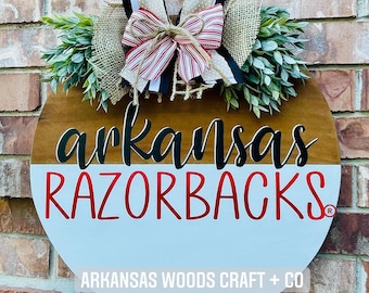May include: A round wooden door hanger with a split design. The top half is brown wood with the words "arkansas RAZORBACKS" in black and red lettering. The bottom half is light blue with the text "ARKANSAS WOODS CRAFT + CO" in black lettering. The door hanger is decorated with a burlap bow, a red and white striped bow, and green leaves.