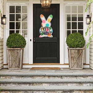 May include: A wooden Easter bunny decoration with colorful polka dots and the words "Happy Easter" hangs on a black front door. The door is framed by white trim and flanked by two windows and matching planters.