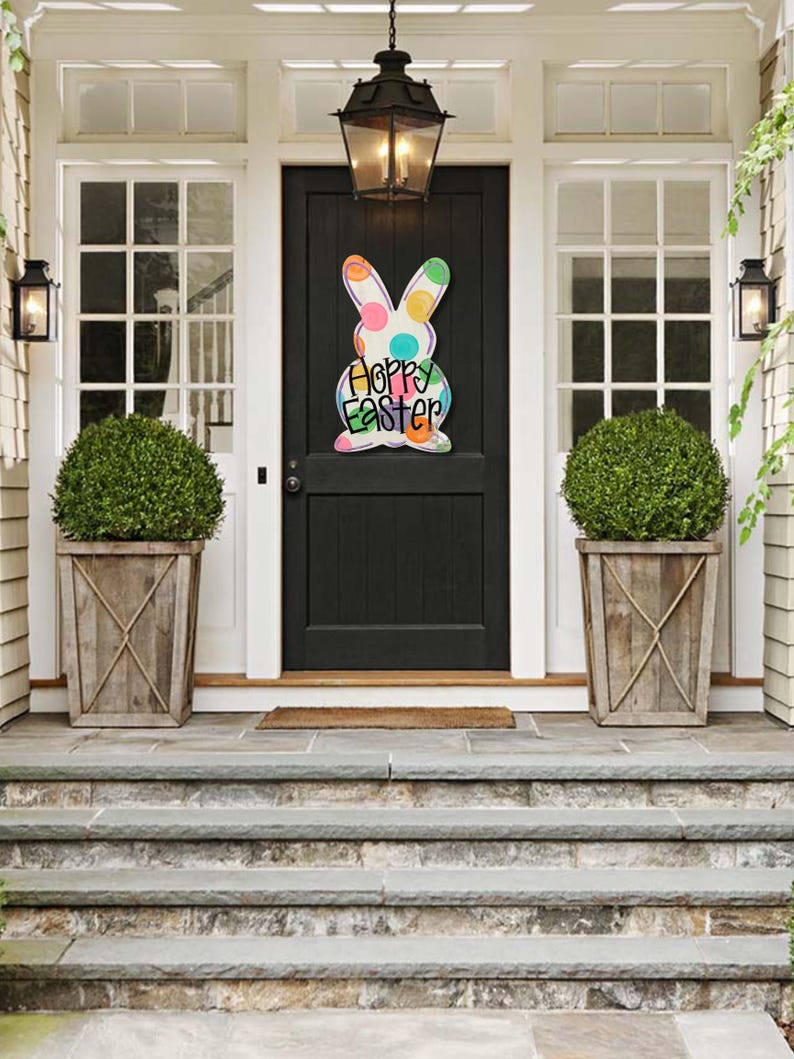 May include: A wooden Easter bunny decoration with colorful polka dots and the words "Happy Easter" hangs on a black front door. The door is framed by white trim and flanked by two windows and matching planters.