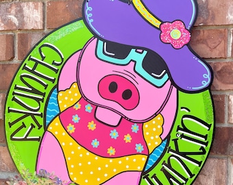 Peut inclure: Une pancarte ronde en bois verte et blanche avec le texte "Summer Fun" écrit en noir. Un cochon rose portant un chapeau violet, des lunettes de soleil et un maillot de bain jaune et rose est au centre de la pancarte.