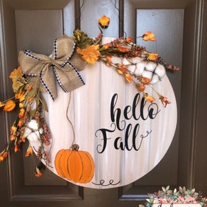 May include: A round wooden door hanger with a whitewashed background and the text "hello fall" in black script. The hanger is decorated with a burlap bow, faux fall leaves, cotton bolls, and a small orange pumpkin.