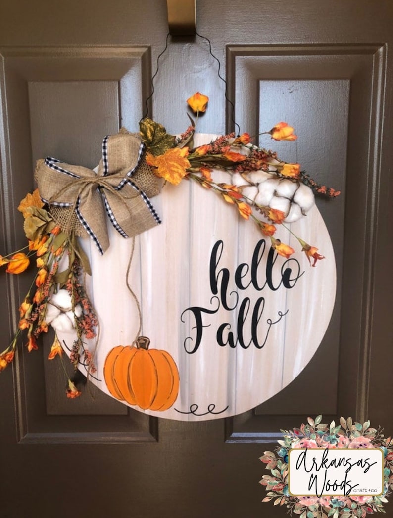 May include: A round wooden door hanger with a whitewashed background and the text "hello fall" in black script. The hanger is decorated with a burlap bow, faux fall leaves, cotton bolls, and a small orange pumpkin.