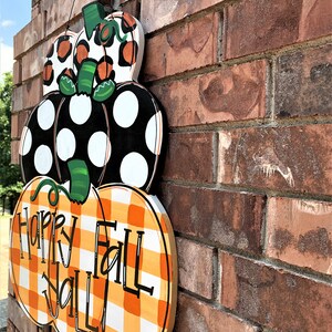 Peut inclure: Une d&eacute;coration de porte en bois en forme de citrouille avec trois citrouilles empil&eacute;es. La citrouille du haut est orange et noire &agrave; imprim&eacute; l&eacute;opard, la citrouille du milieu est noire &agrave; pois blancs et la citrouille du bas est orange et blanche &agrave; carreaux. Les citrouilles ont des tiges vertes et la citrouille du bas porte l'inscription "Happy Fall Y'all!" en noir.