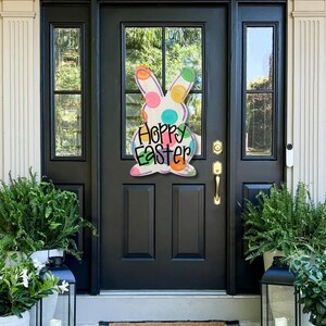 May include: A colorful Easter bunny door hanger with the words "Happy Easter" on a black front door. The bunny has polka dots in various colors. Ferns and lanterns flank the door, creating a welcoming spring scene.