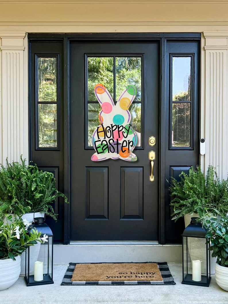 May include: A colorful Easter bunny door hanger with the words "Happy Easter" on a black front door. The bunny has polka dots in various colors. Ferns and lanterns flank the door, creating a welcoming spring scene.