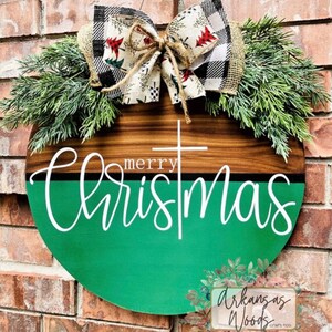 May include: A round wooden sign with a green and brown background. The sign says "merry christmas" in white lettering. The sign is decorated with a burlap bow and greenery.