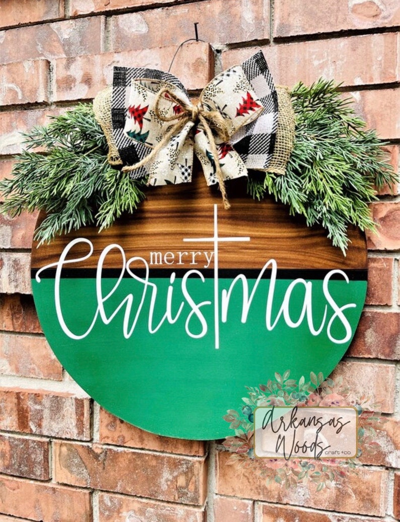 May include: A round wooden sign with a green and brown background. The sign says "merry christmas" in white lettering. The sign is decorated with a burlap bow and greenery.