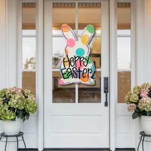 May include: A colorful Easter bunny door hanger with the words "Happy Easter". The bunny is white with polka dots and colorful accents. The door is white with glass panels, and the scene includes potted plants.