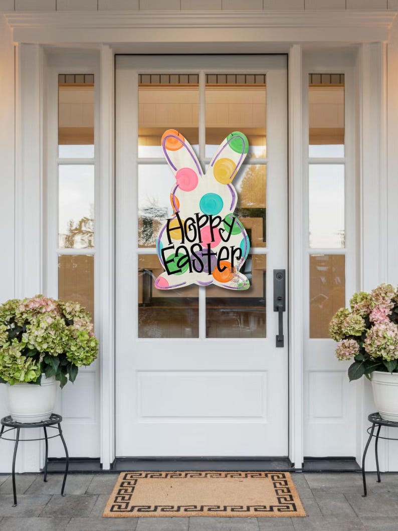 May include: A colorful Easter bunny door hanger with the words "Happy Easter". The bunny is white with polka dots and colorful accents. The door is white with glass panels, and the scene includes potted plants.