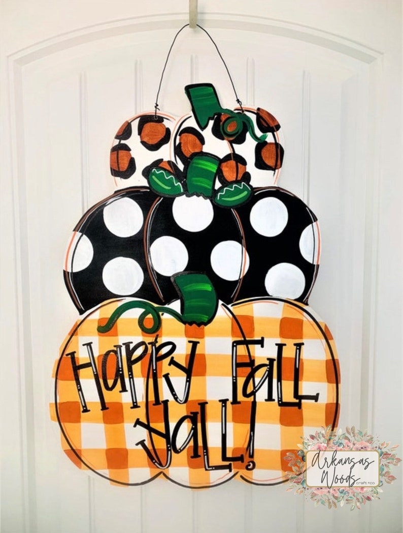 Peut inclure: Une pile de trois citrouilles en bois d&eacute;cor&eacute;es pour l'automne. La citrouille du bas est &agrave; carreaux orange et blanc avec les mots "Happy Fall Y'all!" La citrouille du milieu est noire avec des pois blancs. La citrouille du haut est blanche avec un imprim&eacute; l&eacute;opard noir et orange.