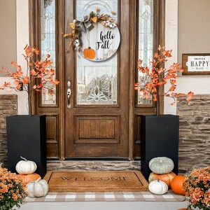 May include: A wooden front door wreath with a white background and the text "hello fall" in black lettering. The wreath is decorated with fall-themed elements, including pumpkins, leaves, and a bow. The wreath is hanging on a brown wooden door with a welcome mat in front of it. There are two black planters with pumpkins on either side of the door.