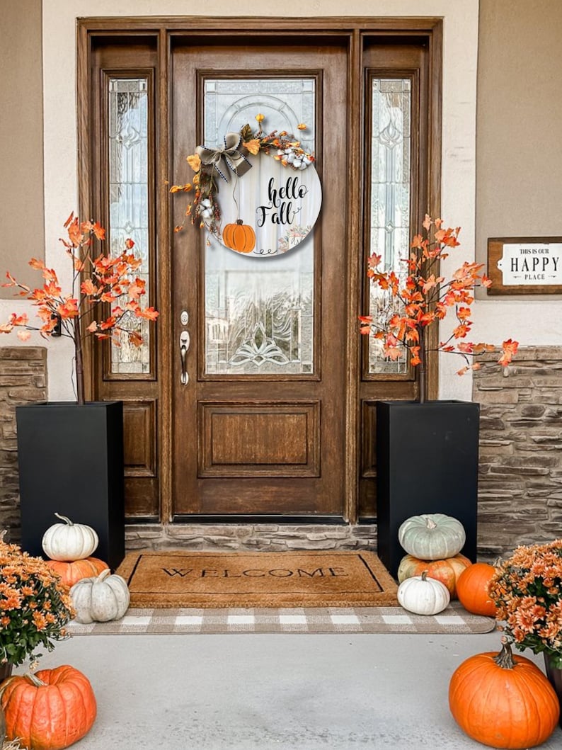 May include: A wooden front door wreath with a white background and the text "hello fall" in black lettering. The wreath is decorated with fall-themed elements, including pumpkins, leaves, and a bow. The wreath is hanging on a brown wooden door with a welcome mat in front of it. There are two black planters with pumpkins on either side of the door.