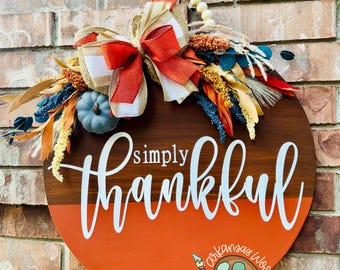 May include: Round wooden sign with the words "simply thankful" in white script. The sign has a brown top half and an orange bottom half. It is decorated with a bow, a small pumpkin, and fall foliage.