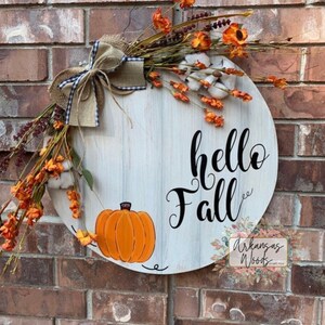 May include: A white wooden round door hanger with the text "hello fall" in black script. The door hanger is decorated with a burlap bow, orange and white flowers, and a painted orange pumpkin.